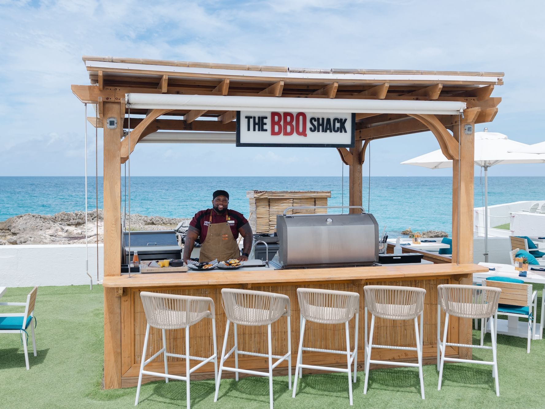 The BBQ Shack, featuring a wooden bar with stools and a chef by the grill facing the ocean at The Morgan Resort Spa Village